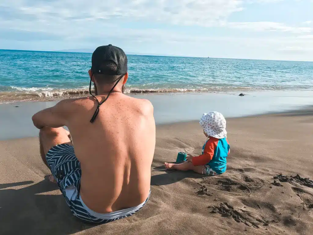 A dad and toddler sitting on a beach, looking out at the ocean in Maui, Hawaii. 