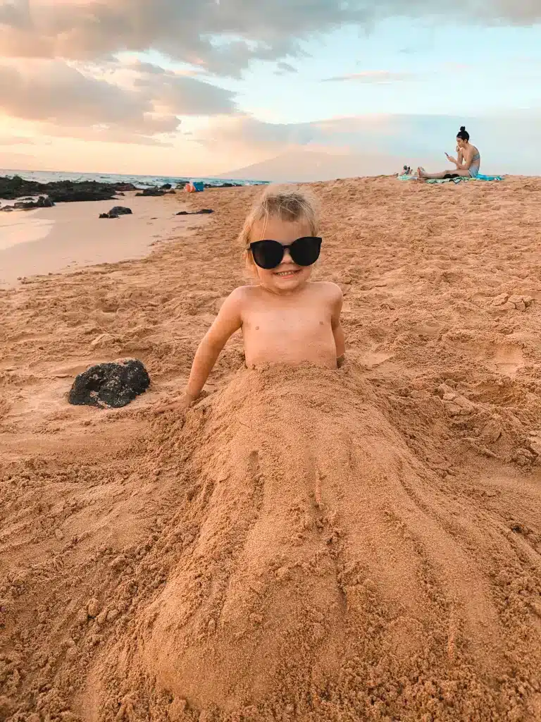 A small child mostly covered in sand and wearing sunglasses on beach in Maui, Hawaii. 