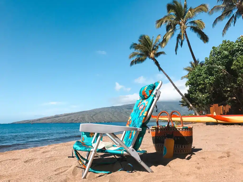 A beach chair and basket on a beach in Hawaii with a canoe in the background. 