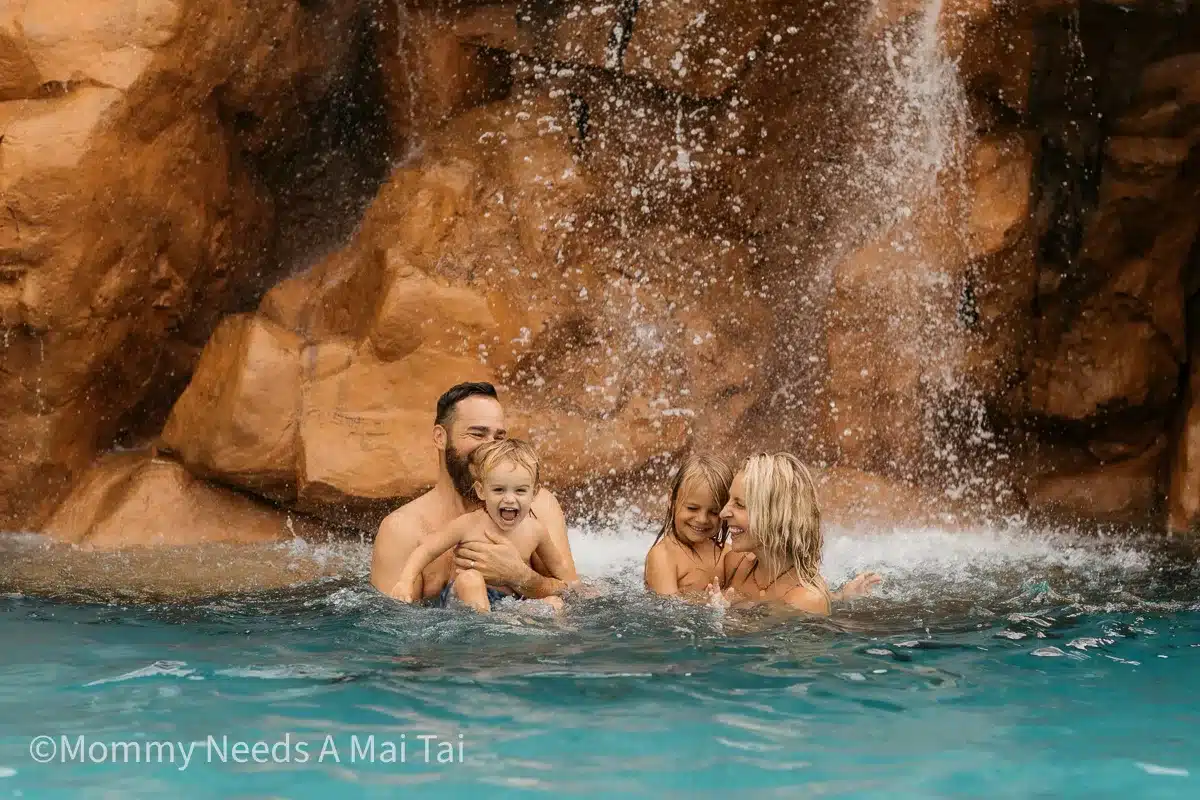 A family with young children playing in a waterfall pool in Hawaii, surrounded by red rock cliffs and tropical scenery.