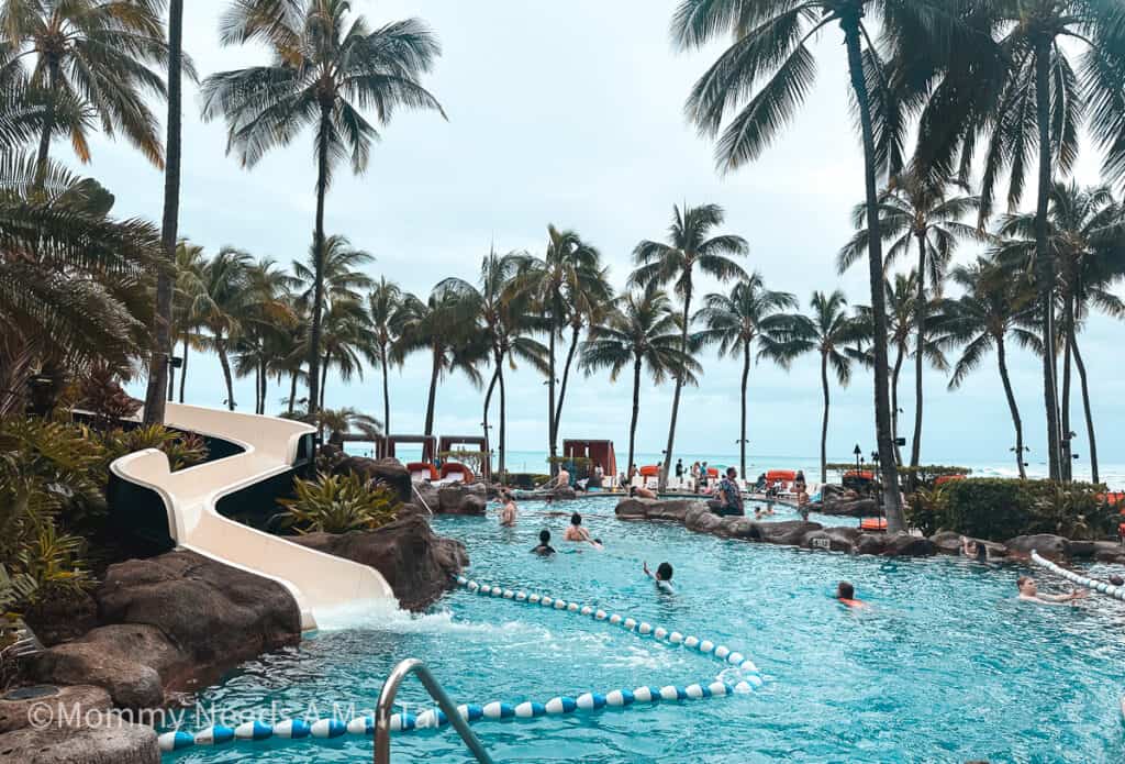 Family pool with waterslide at Sheraton Waikiki, with palm trees and Waikiki Beach in the background.