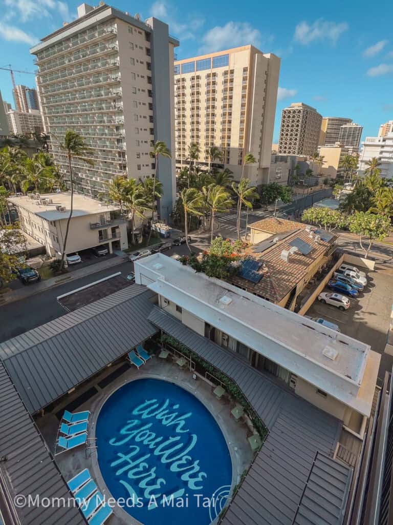 Aerial view of the Surfjack Hotel & Swim Club pool in Waikiki, featuring the iconic “Wish You Were Here” lettering, surrounded by lounge chairs and city buildings.