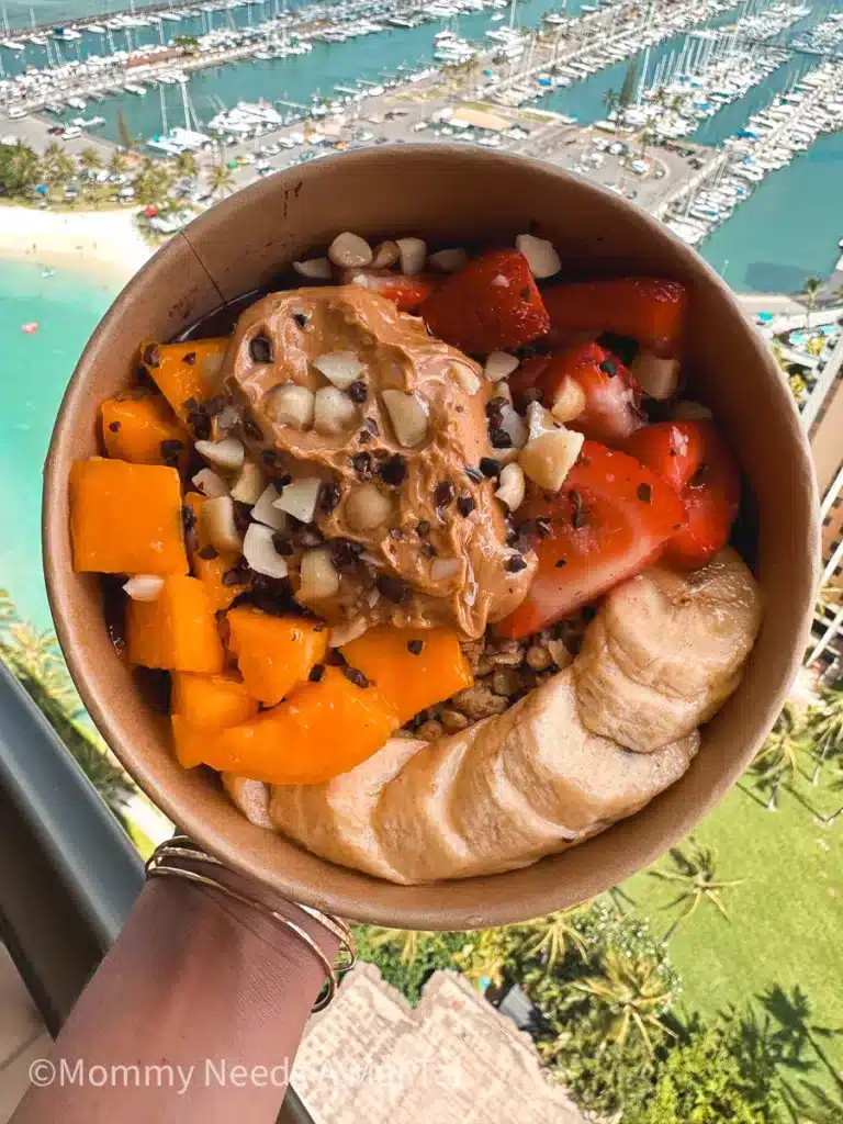 A beauifully decordated acai bowl from Goofy Cafe in Oahu, Hawaii. A hand holds the acai bowl off a high balcony with sailboats in the background.