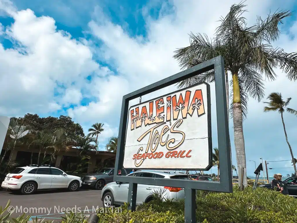 The outside of Haeliwa Joe's Seafood Grill on Oahu, Hawaii, with cars parked and palm trees in the background. 