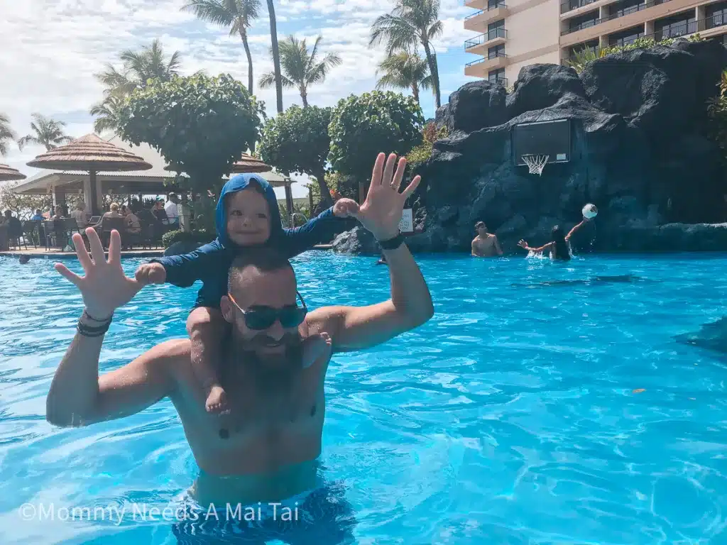 A dad wearing his toddler on his shoulders at a resort pool with people playing basketball in the background. 
