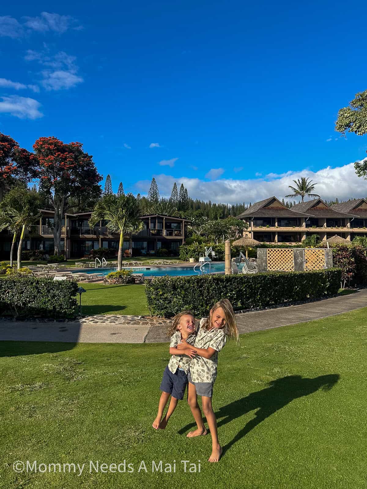 Two kids laughing and playing on the grassy lawn at Napili Kai Beach Resort in West Maui, with the resort buildings and palm trees behind them.