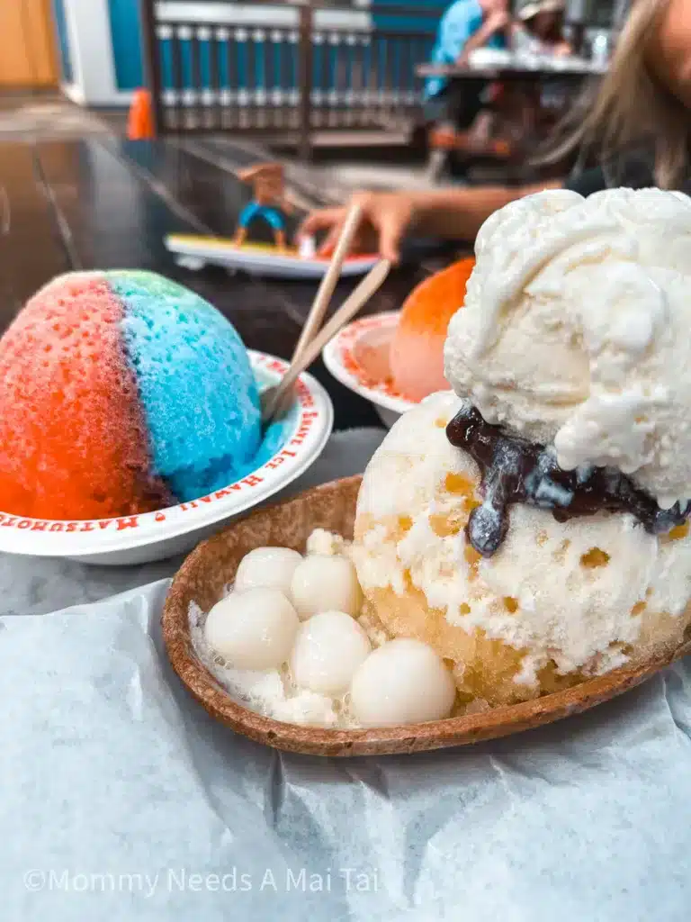 Three different colorful shave ice variations from Motsumoto's Shave Ice on North Shore, Oahu. A kid plays with a toy surfboard in the background. 