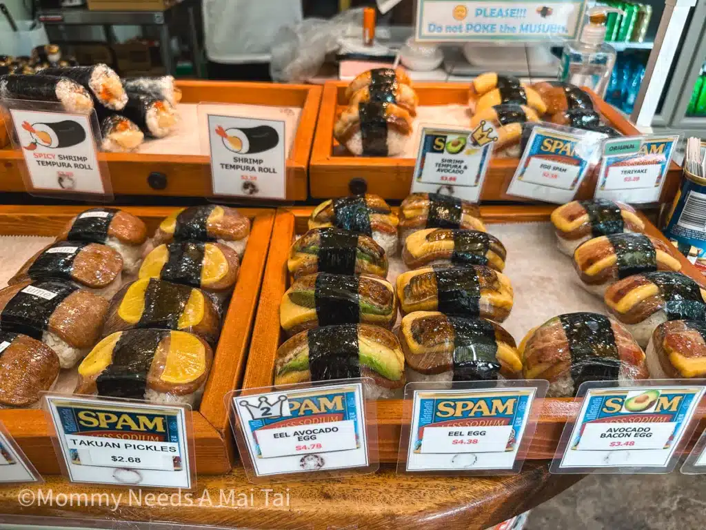 A large selection of Musubis with signs that read "Takuan Pickles," "Eeel Avocado Egg," "Eel Egg," and "Avocado Bacon Egg" at Musiubi Cafe Iyasume in Waikiki, Oahu. 