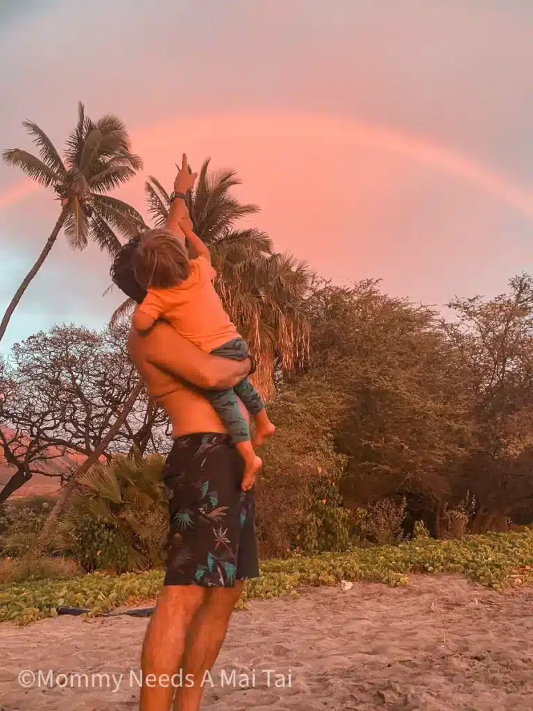 A dad pointing up at a rainbow while holding his toddler in Maui, Hawaii. 