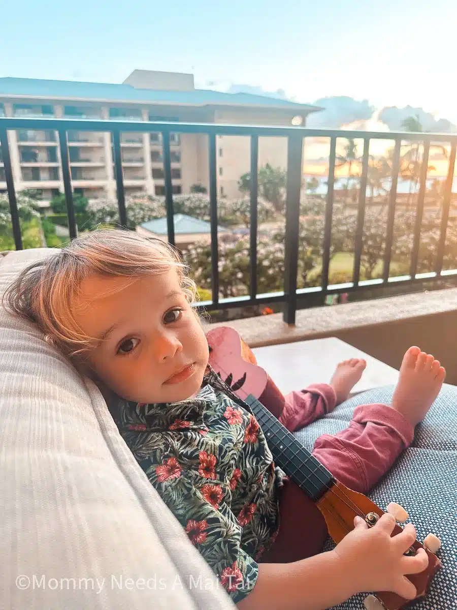A toddler lounging with a ukulele on a lanai at the Four Seasons Resort Maui, overlooking the sunset.