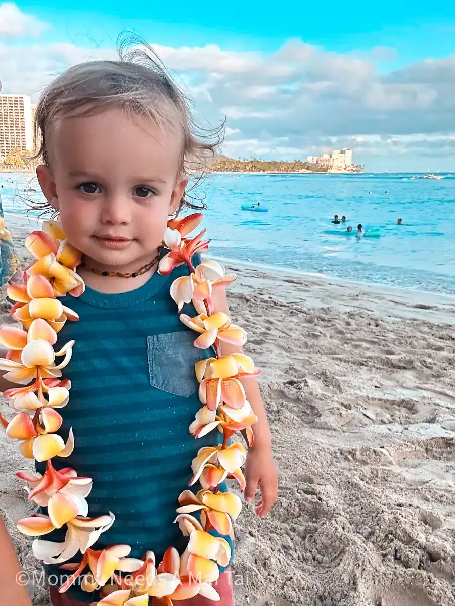 A toddler wearing a flower lei stands on Waikiki Beach with calm blue water and swimmers in the background.