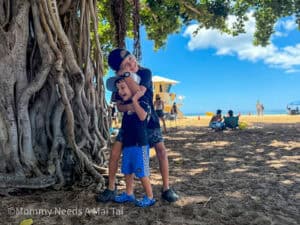 Two young brothers laughing and hugging beneath a large banyan tree on Waikiki Beach, with the ocean, lifeguard tower, and beachgoers in the background.