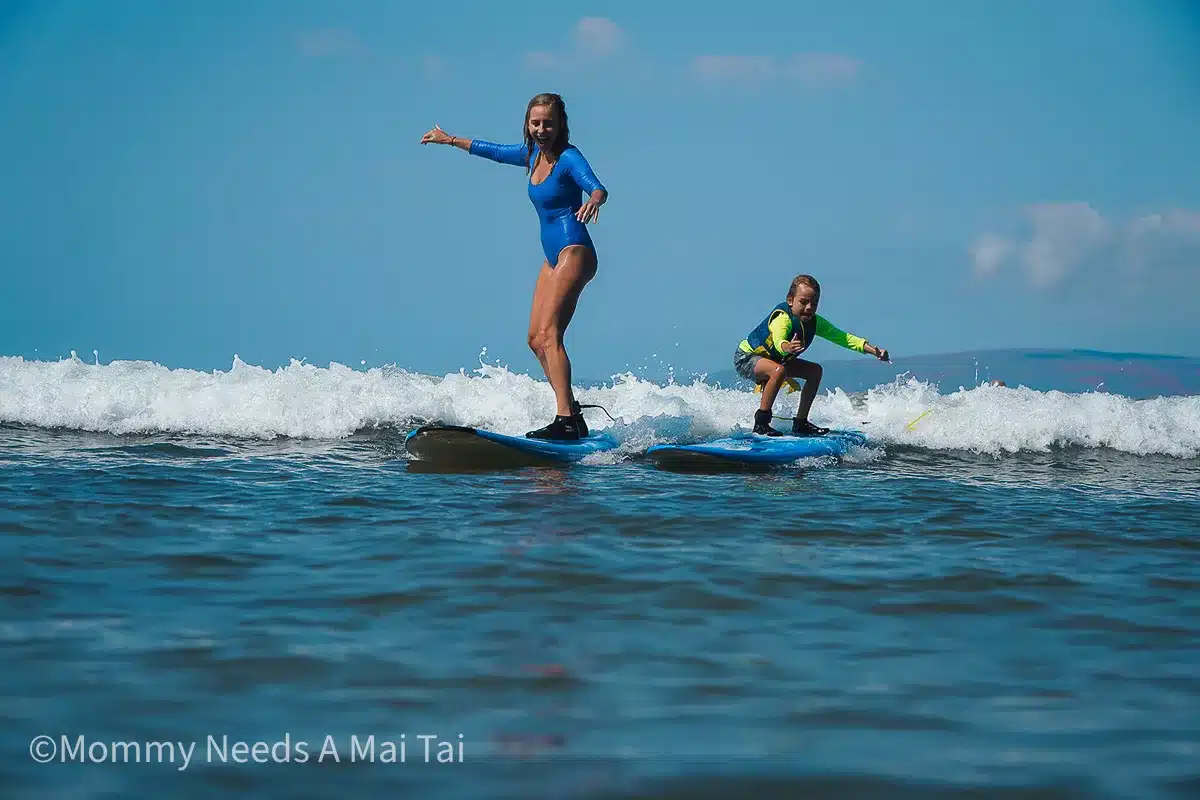 A mom and child looking excited  and nervous as they learn to surf together on small waves in Hawaii.