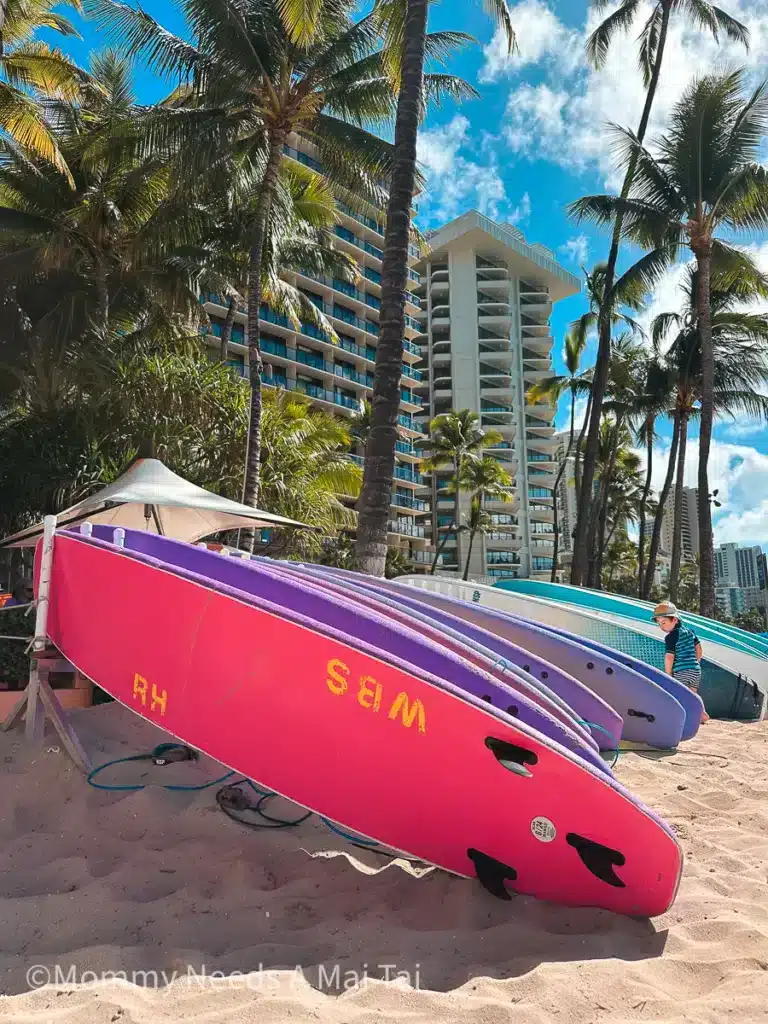 A toddler in a hat standing next to colorful surfboards on the beach in Waikiki, Oahu.