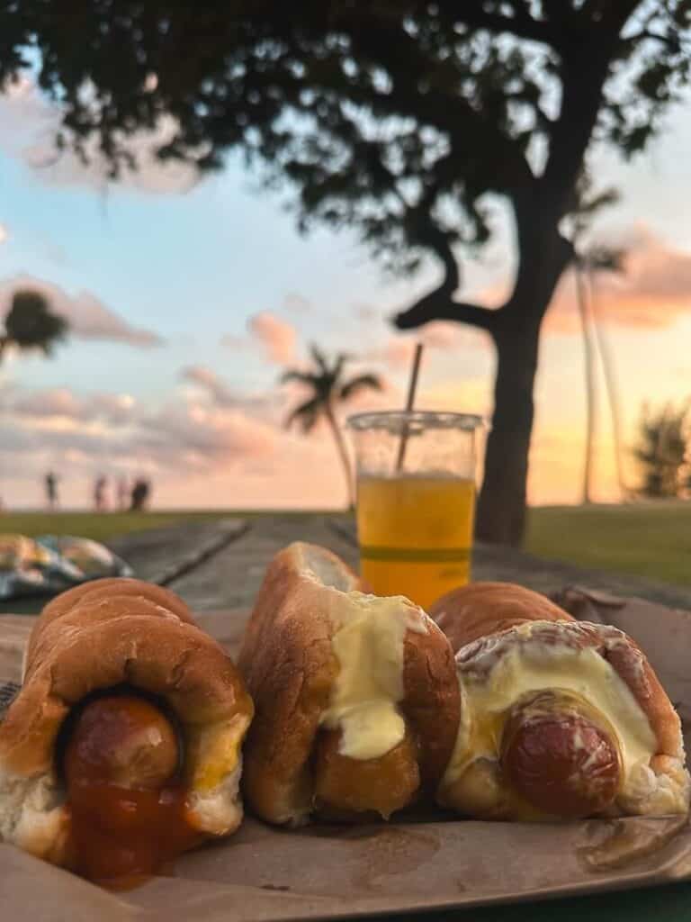 Three Puka Dogs drenched in toppings, next to a lemonade with a sunset in the background at Poipu Beach Park on Kauai.