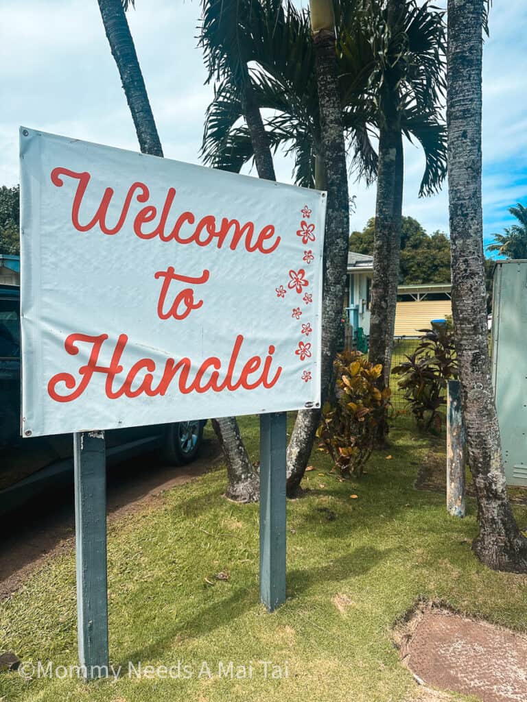 A red and white sign that reads "Welcome to Hanalei" with trees in the background.