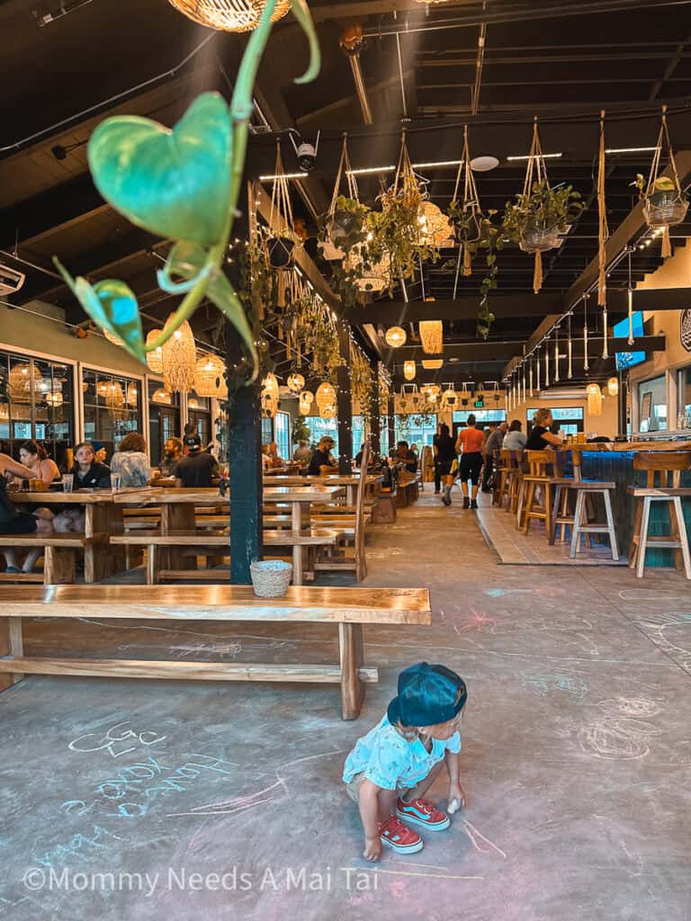 A toddler draws on the floor with chalk and plants hang from the ceiling at Napali Brewing Co on Kauai.