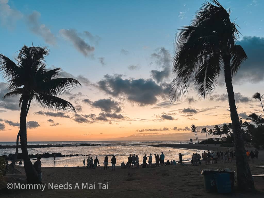 An orange and blue sunset with silhouettes of people and palm trees at Poipu Beach Park on Kauai.