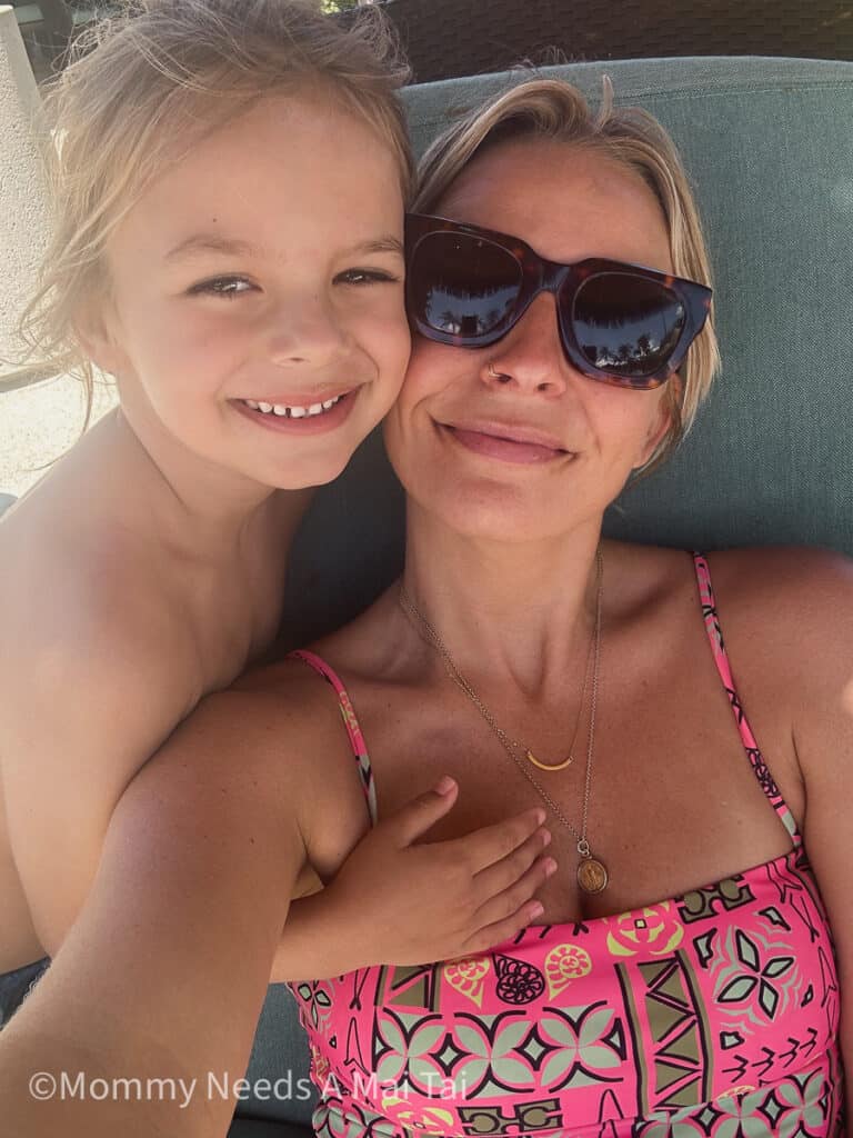 A young boy with long hair smiling and putting his hand on his mom, who is wearing sunglasses and lounging on chair at a resort pool.