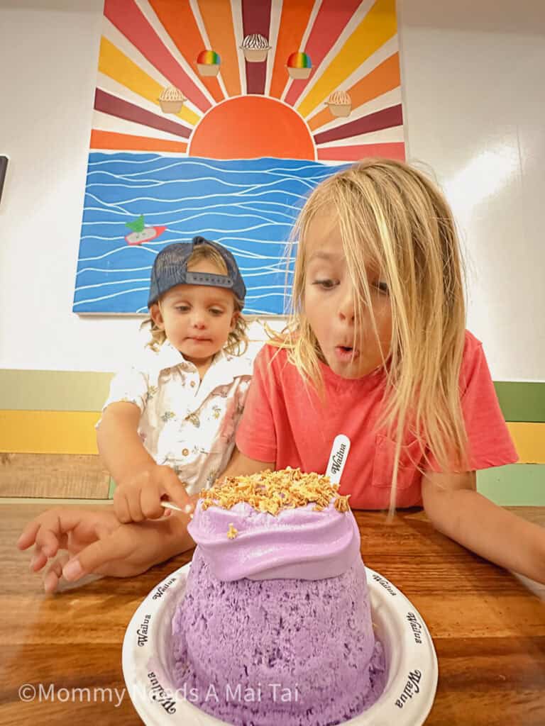 Two kids making excited faces as they look down at a purple shave ice with coconut toppings from Wailua Shave Ice on Kauai.