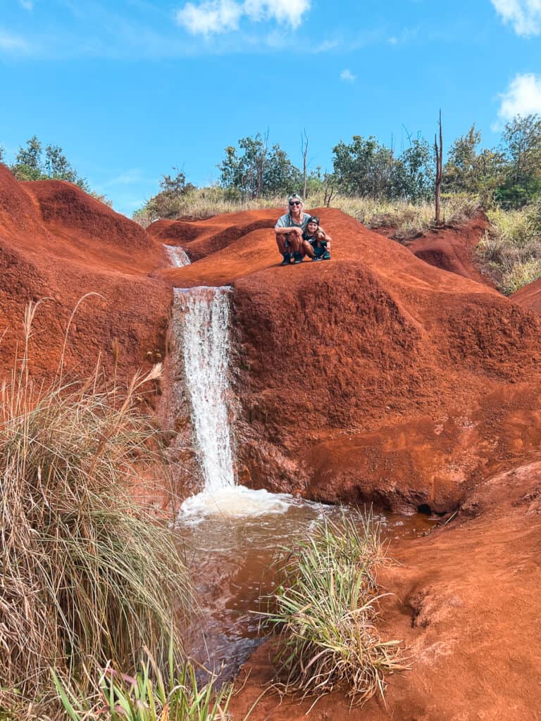 A mom and young boy sitting on top of a Red Dirt Falls mount next to a waterfall in Waimea Canyon, Kauai.