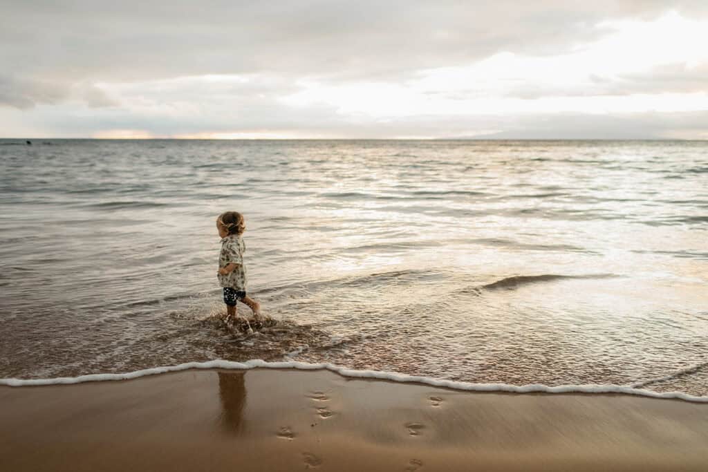A toddler dressed up and walking through the water on a beach in Hawaii. 